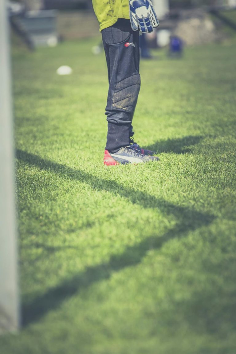 Goalkeeper standing on green field, focused on legs with gear, during a sunny day soccer match.